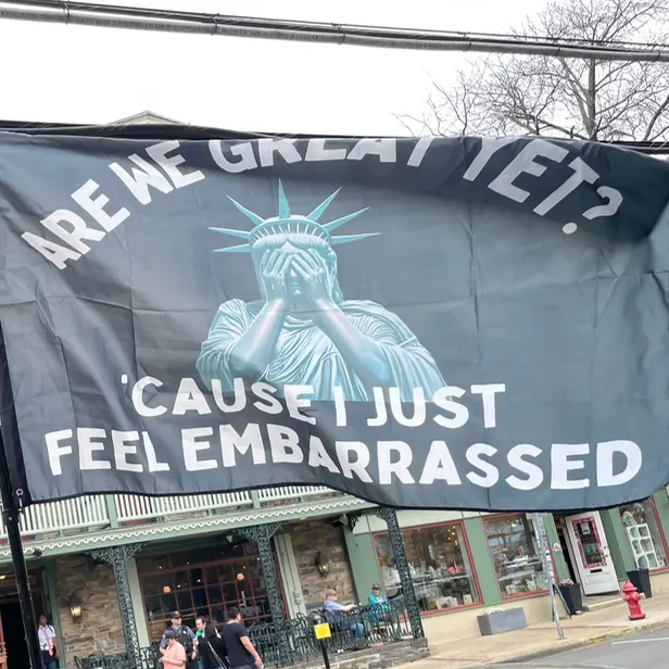 a flag shows the statue of liberty with her head in her hands. the text says are we great yet? &rsquo;cause I just feel embarrassed