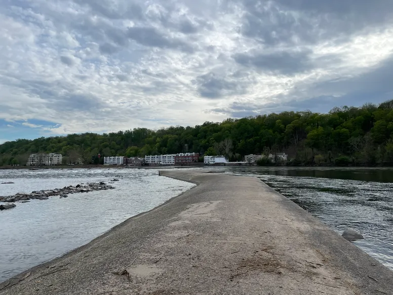 a small concrete dam is underfoot with river water on either side and trees and buildings in the distance