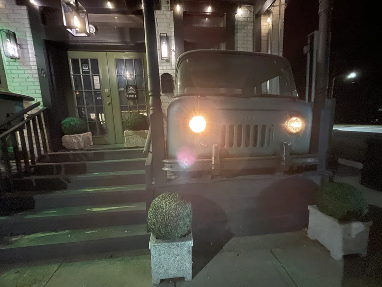 an old Jeep with glowing headlights sits on the porch of a small decortive shop