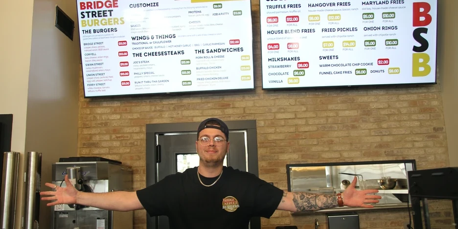 Joe Crozier stands with arms outstretched in front of the menu at the front counter of Bridge Street Burgers