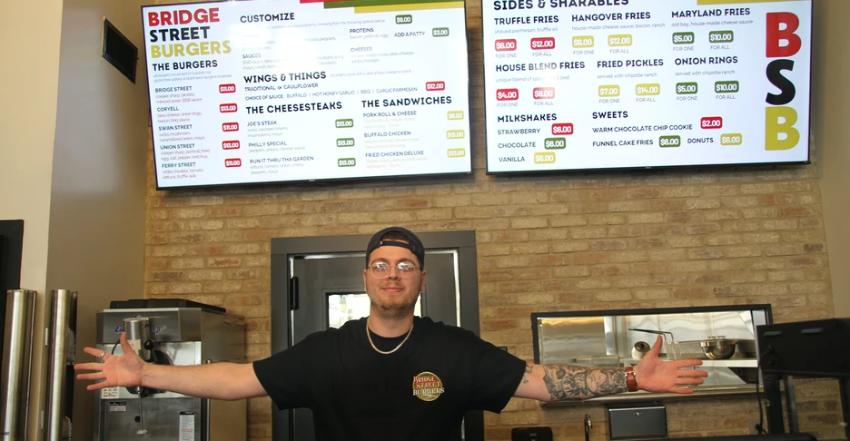 Joe Crozier stands with arms outstretched in front of the menu at the front counter of Bridge Street Burgers