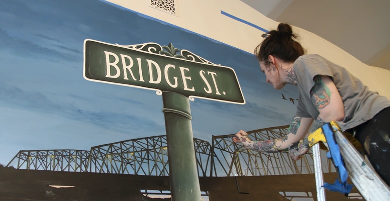 Hope Gaburo stands on a ladder and hand paints a wall mural inside Bridge Street Burgers
