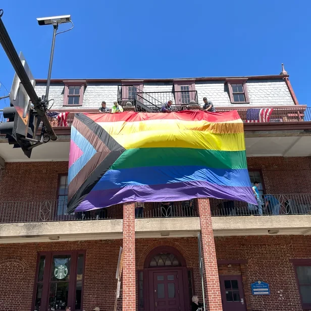 a large progress pride flag hangs over the balcony on the third floor of a building