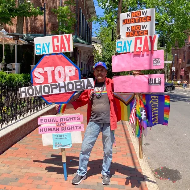 a man holding two elaborate hand signs that read, "say gay, stop homophobia, equal rights are human rights, kNOw justice kNOw peace, equality, act up"