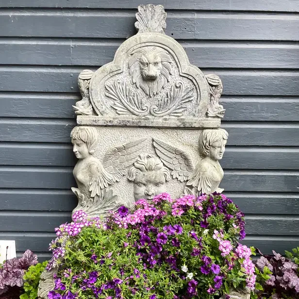 a concrete fountain with a lion and angels carved into the back and what would be the pool of water is instead filled with flowers