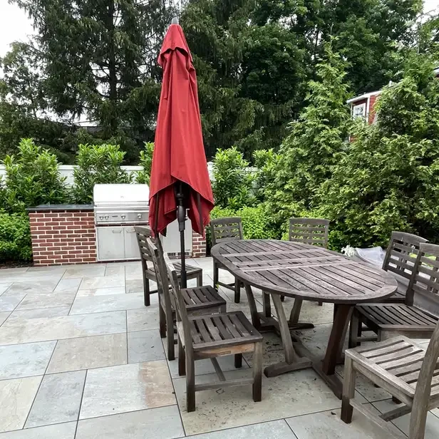 a patchwork of stone slates form a patio with a wood table, umbrella, and grill on top