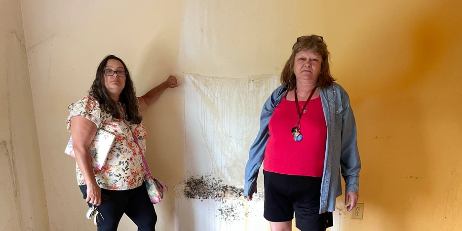 two frowning women stand by a stained wall that has black mold growing on it