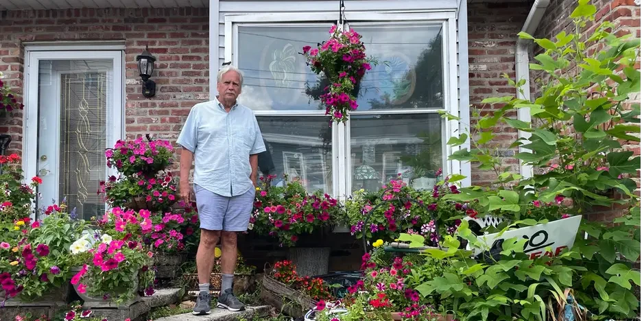 a man with white hair stands on his front porch, surrounded by flowers