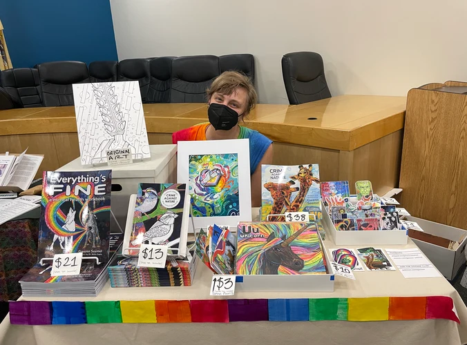 Gwenn sitting behind a table that has books, stickers, prints, cards, and more