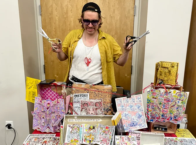 A. T. Pratt poses with two pairs of scizzors behind his table, which is covered with his handmade comic zines