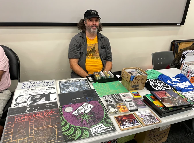 T sitting behind his table, which has LP records, CDs, cassette tapes, and t-shirts for sale