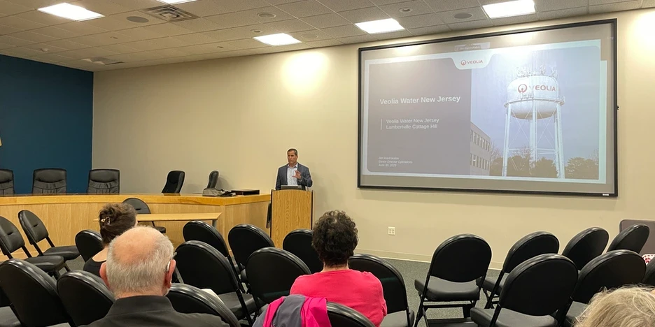 a man wearing a suit without a tie stands at a poduim beside a slide presentation and speaks to a small, in-person crowd