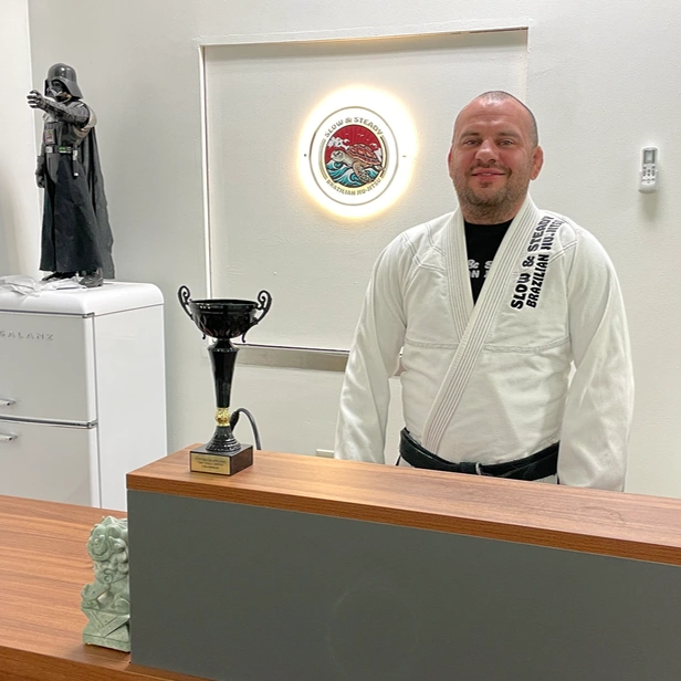 Nick smiles as he stands behind the desk at his new gym, proudly wearing a gi and blackbelt. a Darth Vader statue and ‘Slow and Steady’ logo light his behind him.