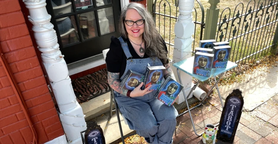 Kristine, with long, wavey gray hair and wearing her signature overalls, smiles as she sits holding several Tarot decks