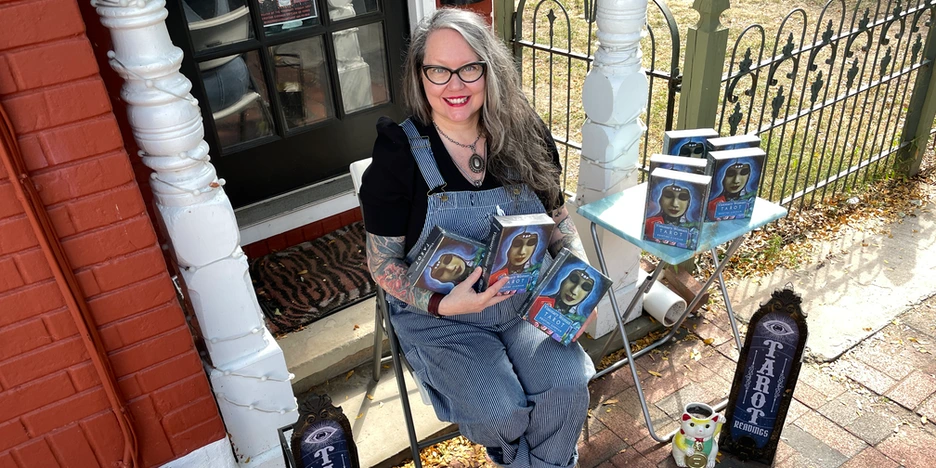 Kristine, with long, wavey gray hair and wearing her signature overalls, smiles as she sits holding several Tarot decks