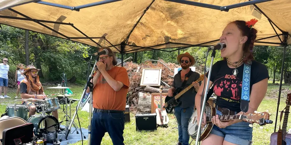 outdoors, a funky woman holds a banjo and sings into a microphone. behind her is a bassist, drummer, and harmonica player.