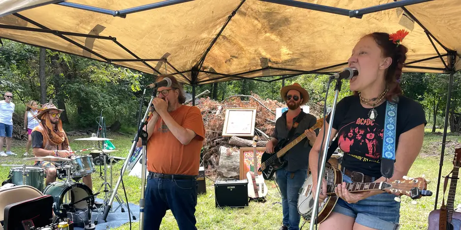 outdoors, a funky woman holds a banjo and sings into a microphone. behind her is a bassist, drummer, and harmonica player.