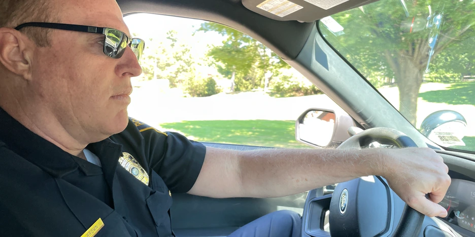 Robert Brown, a police officer, wearing sunglasses, drives a police cruiser with his hand on the steering wheel